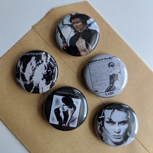 Peut inclure: Ensemble de cinq badges bouton noirs et blancs représentant des images d'Adam Ant. Les badges présentent des pochettes d'album des albums "Dirk Wears White Sox", "Friend or Foe", "Strip", "Friend or Foe" et "Lady".