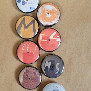 Puede incluir: Ocho botones redondos y coloridos con bordes negros. Los botones tienen varios diseños, incluyendo un botón blanco con un punto negro en el centro, un botón rojo con texto blanco, un botón negro con texto blanco, un botón marrón con un punto negro en el centro, un botón amarillo con texto negro y un botón azul con texto negro.