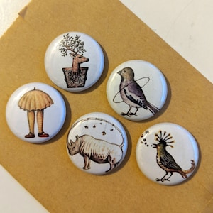 May include: Set of six round pinback buttons featuring whimsical illustrations. The buttons depict a deer in a pot, a bird with a ring around it, a person with an umbrella, a rhinoceros, a bird with a crown, and a bird with a long tail.
