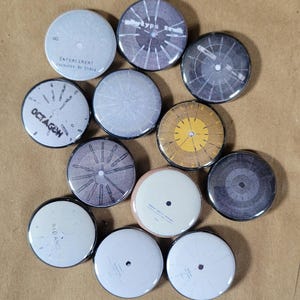 Puede incluir: Una colección de 12 botones redondos con borde negro y varios diseños. Los botones presentan patrones circulares en blanco y negro, algunos con texto, incluyendo "OCTAGON", "ENFORCEMENT", "BAD" y "METRO TYPE".