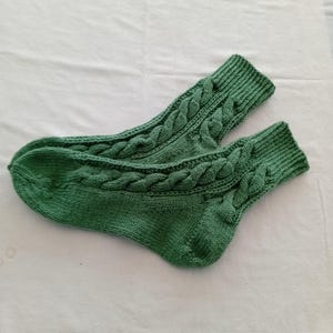 Könnte beinhalten: Ein Paar grüne Stricksocken mit Zopfmuster. Die Socken sind einfarbig grün und scheinen handgefertigt zu sein. Das Zopfmuster verläuft über den Fuß und den Schaft der Socke.