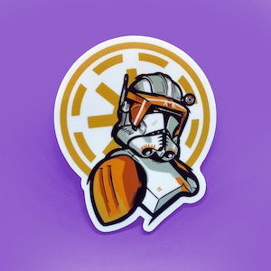 May include: A sticker featuring a cartoon illustration of a Star Wars clone trooper wearing an orange and white helmet and armor. The trooper is standing in front of a gold and white symbol.