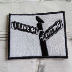Peut inclure: Patch brodé blanc avec des points noirs. Le patch représente un panneau de rue qui indique "I Live In East Van".