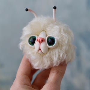 May include: A handmade, fluffy white creature with large, expressive green eyes and a pink nose. The whimsical design includes two small antennae with black tips. This unique art piece is held in a hand against a soft blue background.