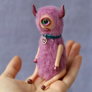 May include: A pink, fuzzy, one-eyed creature with horns sits on a hand. The creature has a teal blue collar with a silver peace sign charm.
