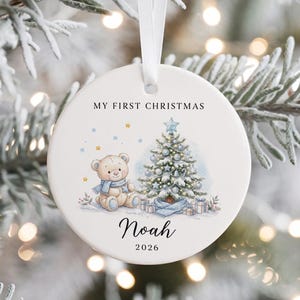 Op de afbeelding: Een ronde witte keramische kerstbal met een wit lint. Het ontwerp toont een teddybeer met een blauwe sjaal, een versierde kerstboom en de tekst "MY FIRST CHRISTMAS", "Noah" en "2026" in zwarte letters.