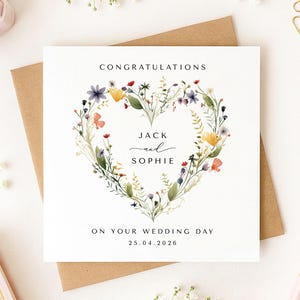 May include: A wedding card featuring a floral heart design. The card is inscribed with "CONGRATULATIONS", the names "JACK and SOPHIE", and the date "25.04.2026". The card is placed on a brown envelope, with floral accents.