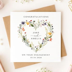 May include: A white engagement card with a floral heart design. The card reads "CONGRATULATIONS" at the top, with the names "JAKE and AMELIA" in the center, and "ON YOUR ENGAGEMENT 30.04.2026" at the bottom. The card is on a brown envelope.