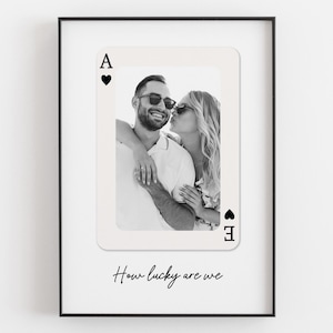 May include: A framed wall print featuring a custom playing card design with an Ace of Hearts symbol. The centre displays a black and white photograph of two people embracing. Below the card, the text reads, How lucky are we, in black script.
