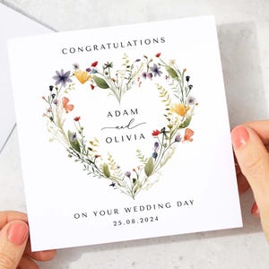 May include: A white wedding congratulations card with a floral heart design. The card features the names "Adam and Olivia" and the date "25.08.2024". The floral heart includes colorful wildflowers and greenery, with the word "Congratulations" at the top.