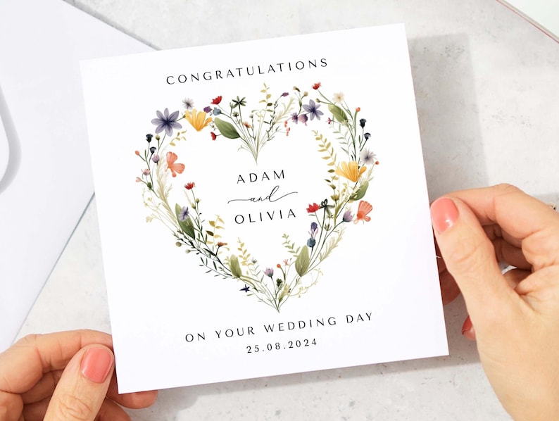May include: A white wedding congratulations card with a floral heart design. The card features the names "Adam and Olivia" and the date "25.08.2024". The floral heart includes colorful wildflowers and greenery, with the word "Congratulations" at the top.