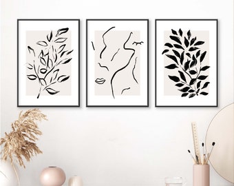 May include: Three framed art prints with black frames. The prints feature minimalist black line drawings on a beige background. One print shows a face, and the other two feature botanical designs of leaves and branches. The artwork is displayed on a white surface.