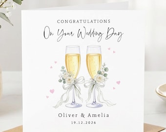 May include: A square white wedding greeting card featuring the text CONGRATULATIONS On Your Wedding Day, Oliver & Amelia, and 19.12.2026. The design includes two champagne flutes with yellow liquid, white floral accents, and ribbons.