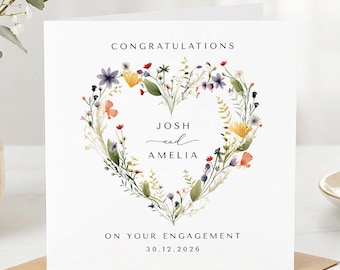 May include: A square white greeting card featuring a heart-shaped wreath made of colourful wildflowers. The text reads CONGRATULATIONS, JOSH and AMELIA, ON YOUR ENGAGEMENT, and 30.12.2026. The card is displayed against a plain white background.
