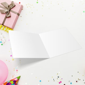 May include: An open, blank greeting card on a white surface, surrounded by party decorations. A pink gift box with a brown ribbon, a pink balloon, a party hat, and gold star-shaped confetti are visible. Yellow streamers and colorful confetti add to the festive scene.