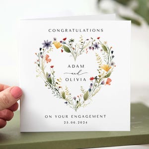 May include: A white engagement card with a floral heart design. The card reads "Congratulations" at the top, with the names "Adam and Olivia" in the center, and "On your engagement 25.06.2024" at the bottom. The floral heart includes purple, yellow, and orange flowers.