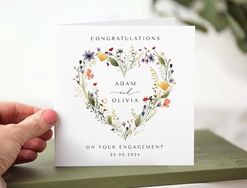May include: A white engagement card with a floral heart design. The card reads "Congratulations" at the top, with the names "Adam and Olivia" in the center, and "On your engagement 25.06.2024" at the bottom. The floral heart includes purple, yellow, and orange flowers.