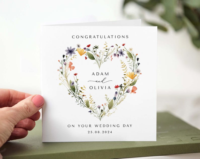 May include: A white wedding card with the words "Congratulations" and "On Your Wedding Day" printed in black. The card features a floral heart design with the names "Adam and Olivia" and the date 25.08.2024.