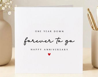 May include: A white anniversary card with black script text: "One Year Down, forever to go, Happy Anniversary." A small red heart is below the text. The card is on a white background.