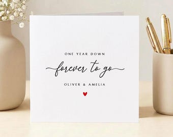 May include: A white square card with the words "ONE YEAR DOWN forever to go" in black script. The names "OLIVER & AMELIA" are below the text, with a small red heart. The card is displayed with a vase of flowers and pens.