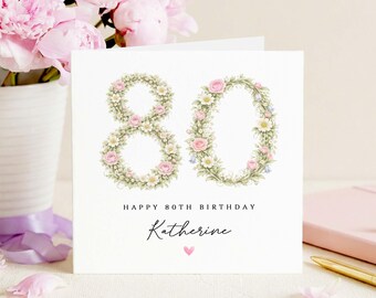 May include: A white birthday card featuring the number 80, crafted from floral wreaths of pink roses and white daisies. The card reads "Happy 80th Birthday" and "Katherine", with a small pink heart. A vase of pink flowers is in the background.