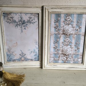 Könnte beinhalten: Zwei gerahmte Kunstdrucke mit weißen, verwitterten Rahmen. Einer zeigt ein Blumendesign auf hellem Hintergrund, der andere ein Blumenmuster auf blau-weißen Streifen. Die Drucke haben eine Vintage-Ästhetik.