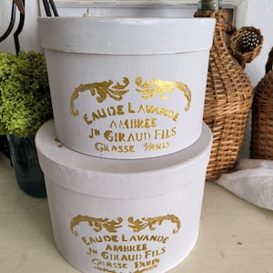 Könnte beinhalten: Zwei gestapelte, runde, weiße Schachteln mit goldfarbenen Buchstaben und Akzenten. Die Schachteln haben einen Deckel und sind mit "EAUDE LAVANDE AMBREE JN GIRAUD FILS GRASSE PARIS" beschriftet. Die Schachteln dienen wahrscheinlich zur Aufbewahrung oder Präsentation.
