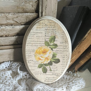 Könnte beinhalten: Ein ovales Holzschild mit einem Vintage-Druck einer gelben Rose mit grünen Blättern, überlagert von einem Hintergrund aus gealterter französischer Schrift und numerischem Buchhaltungstext, mit einem hellen, leicht abgenutzten Rand.