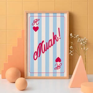 Puede incluir: Una impresión artística enmarcada con un diseño de carta de juego. Presenta rayas verticales azules y blancas, un símbolo de corazón rojo y la palabra "Muak!" en cursiva. El marco es de madera clara.