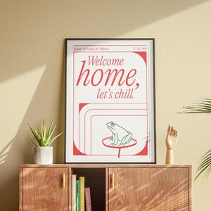 Puede incluir: Una impresión artística enmarcada con las palabras "Welcome home, let's chill" en escritura roja. La impresión presenta una rana verde sobre una hoja de nenúfar. El marco es negro y la impresión se muestra en un mueble de madera.