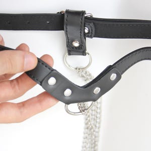 May include: Black leather belt with a silver chain and metal rings. The belt has a buckle and a strap with a D-ring.