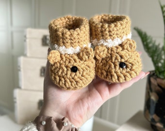 May include: A pair of handmade crochet baby booties in a light brown color, designed to resemble teddy bears. The booties have black button eyes and white trim around the ankles. These soft, cozy booties are perfect for a newborn.