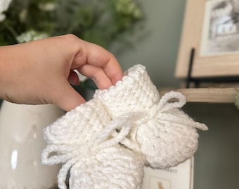 May include: A pair of handmade, off-white crochet baby booties. The booties have a rounded toe and are tied with a matching yarn bow. The soft, textured yarn creates a cozy and delicate appearance, perfect for a newborn.