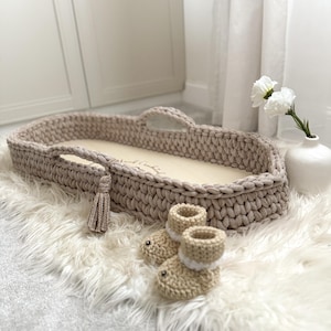 May include: A beige crochet baby bassinet with a wooden base and handles, alongside a pair of matching crochet baby booties. The bassinet has a decorative tassel and is on a fluffy white rug.