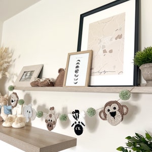 May include: A handmade crochet animal garland featuring a monkey, zebra, giraffe, elephant, and other animals, with green pom-poms. The garland is displayed on a shelf with framed artwork and decorative items. The color palette includes beige, brown, white, and green.