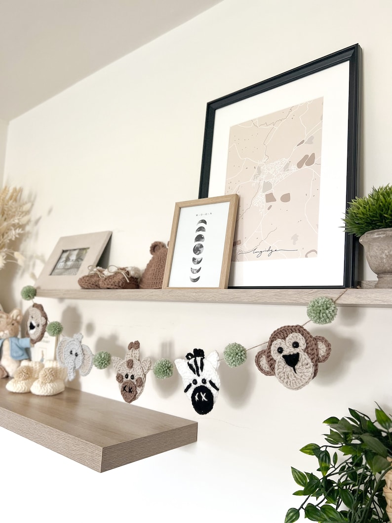 May include: A handmade crochet animal garland featuring a monkey, zebra, giraffe, elephant, and other animals, with green pom-poms. The garland is displayed on a shelf with framed artwork and decorative items. The color palette includes beige, brown, white, and green.