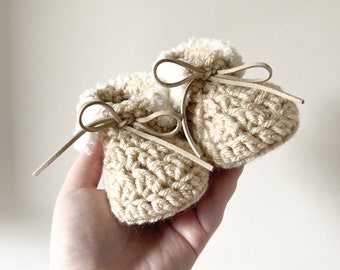 May include: A pair of handmade baby booties in a light beige color. The booties are crocheted with a textured stitch and feature a soft, fluffy lining. Each bootie has a leather tie, adding a touch of elegance. These cozy booties are perfect for a newborn.