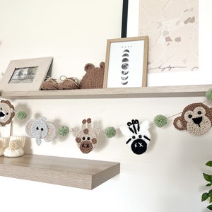 May include: A handmade crochet animal garland featuring a lion, elephant, giraffe, zebra, and monkey. The garland is strung with green pom-poms and is displayed on a shelf with other nursery decor.