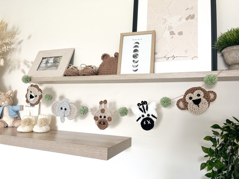 May include: A handmade crochet animal garland featuring a lion, elephant, giraffe, zebra, and monkey. The garland is strung with green pom-poms and is displayed on a shelf with other nursery decor.