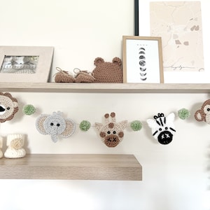 May include: A handmade crochet safari animal garland with a lion, elephant, giraffe, zebra, and monkey. The garland is strung with green pom-poms. Additional items include a pair of baby booties, a bear head, and a framed picture on a wooden shelf.