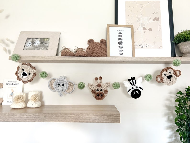 May include: A handmade crochet safari animal garland with a lion, elephant, giraffe, zebra, and monkey. The garland is strung with green pom-poms. Additional items include a pair of baby booties, a bear head, and a framed picture on a wooden shelf.