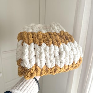 May include: A chunky knit beanie in a striped pattern of white and mustard yellow. The hat is made of thick, soft yarn, and appears to be hand-knitted. The beanie is folded over, showing the interior. The background is a plain white wall.