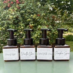 May include: Four brown glass foaming hand soap bottles with black pumps. Each bottle has a white label with text: "Harvest Apple", "Vanilla Chai", "Oatmeal Milk + Honey", and "Pumpkin Spice". The bottles are on a green surface with a blurred green background.