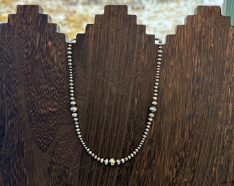 May include: A silver beaded necklace displayed on a dark wooden jewelry stand. The necklace features small, round beads that graduate in size, with larger beads at the center. The stand has a decorative, stepped design.