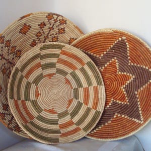 May include: Three round woven wall baskets featuring intricate patterns. The largest basket displays a tan base with orange geometric floral motifs. The center basket has a spiral design in olive green and orange. The third basket features a large star.