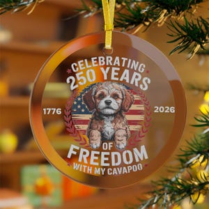 Könnte beinhalten: Runder, klarer Glasornament mit goldenem Band zum Aufhängen. Das Ornament zeigt eine Cavapoo-Hundeillustration mit amerikanischem Flaggenhintergrund und dem Text "Celebrating 250 Years of Freedom with my Cavapoo".