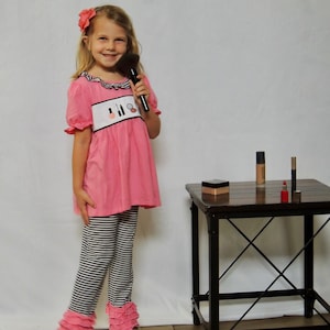 May include: A young girl wearing a pink shirt with makeup icons and black and white striped pants with pink ruffles. She is holding a makeup brush and looking at the camera. There is a small table with makeup products on it.