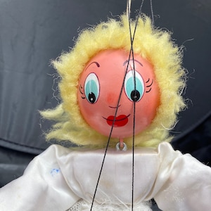 Puede incluir: Un títere de mano vintage con una cara pintada, pelo de lana rubio y un vestido blanco con una superposición de encaje. El títere está unido a cuerdas para su manipulación.