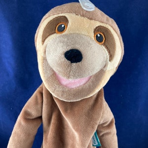 May include: A brown sloth hand puppet with a big smile and large, brown eyes. The puppet is wearing a brown shirt and has a green tag that says "Fiesta" on it.