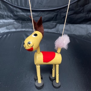 Puede incluir: Un títere de madera de un caballo con una silla de montar roja, una crin marrón y una cola rosa. El títere está en cuerdas y tiene ruedas negras.